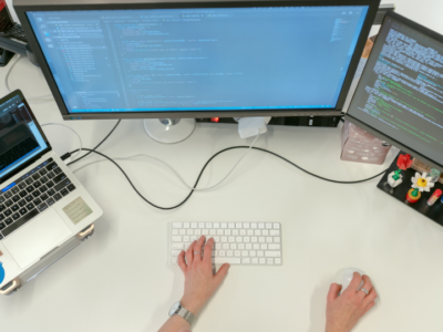 A white desk is shown from above, with, from left to right, an open laptop and two computer screens on it. In the center of the image, we see a person's hands operating a keyboard and mouse.