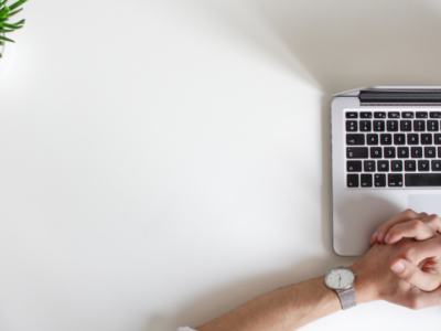 The image is shot from above. It shows a man's hands clasped together over an open Macbook laptop places on a while table. A desk plant peeks in from the top left corner of the image.