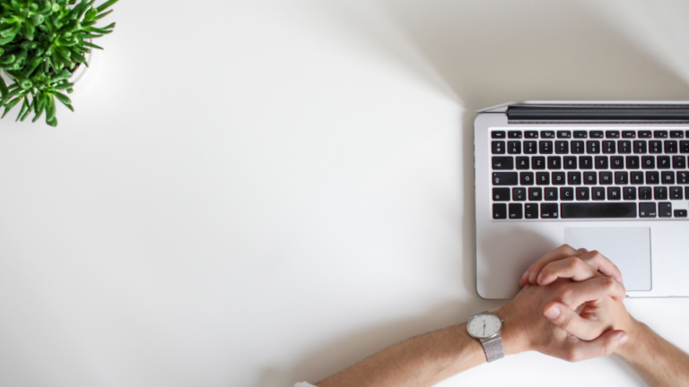 The image is shot from above. It shows a man's hands clasped together over an open Macbook laptop places on a while table. A desk plant peeks in from the top left corner of the image.