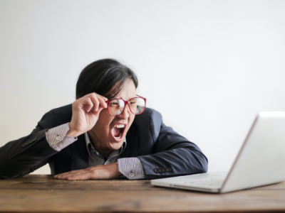 This image shows a guy in a suit and red glasses, with his arms on a wooden table, looking at a laptop with an amazement expressed with an open mouth and while holding his glasses. This photo is attached to a case study about how a multinational bank reduced their hackability by over 50%​