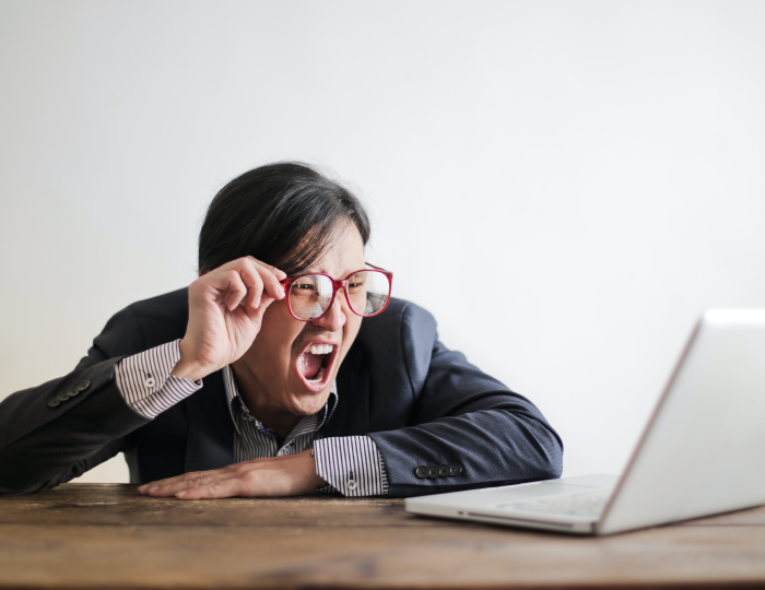 This image shows a guy in a suit and red glasses, with his arms on a wooden table, looking at a laptop with an amazement expressed with an open mouth and while holding his glasses. This photo is attached to a case study about how a multinational bank reduced their hackability by over 50%​